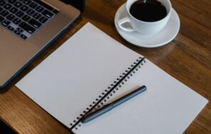 ノートパソコンの横に白紙のメモ帳とコーヒーを置いたデスクのイメージ