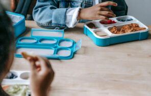 Crop faceless classmates sitting at table with lunch boxes with various food and eating during break