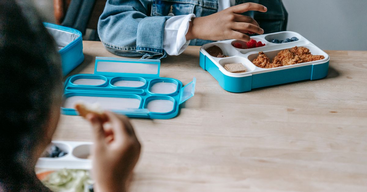 Crop faceless classmates sitting at table with lunch boxes with various food and eating during break